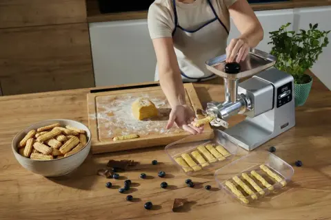 Ręcznie robione ciastka przez maszynkę, świeżo wyciśnięte, obok miska pełna ciastek, jagody i czekolada.