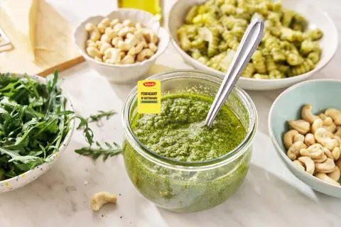 Słoik pełen aromatycznego pesto z rukoli, obok miseczki z orzechami nerkowca i świeżą rukolą. Idealne do makaronu!