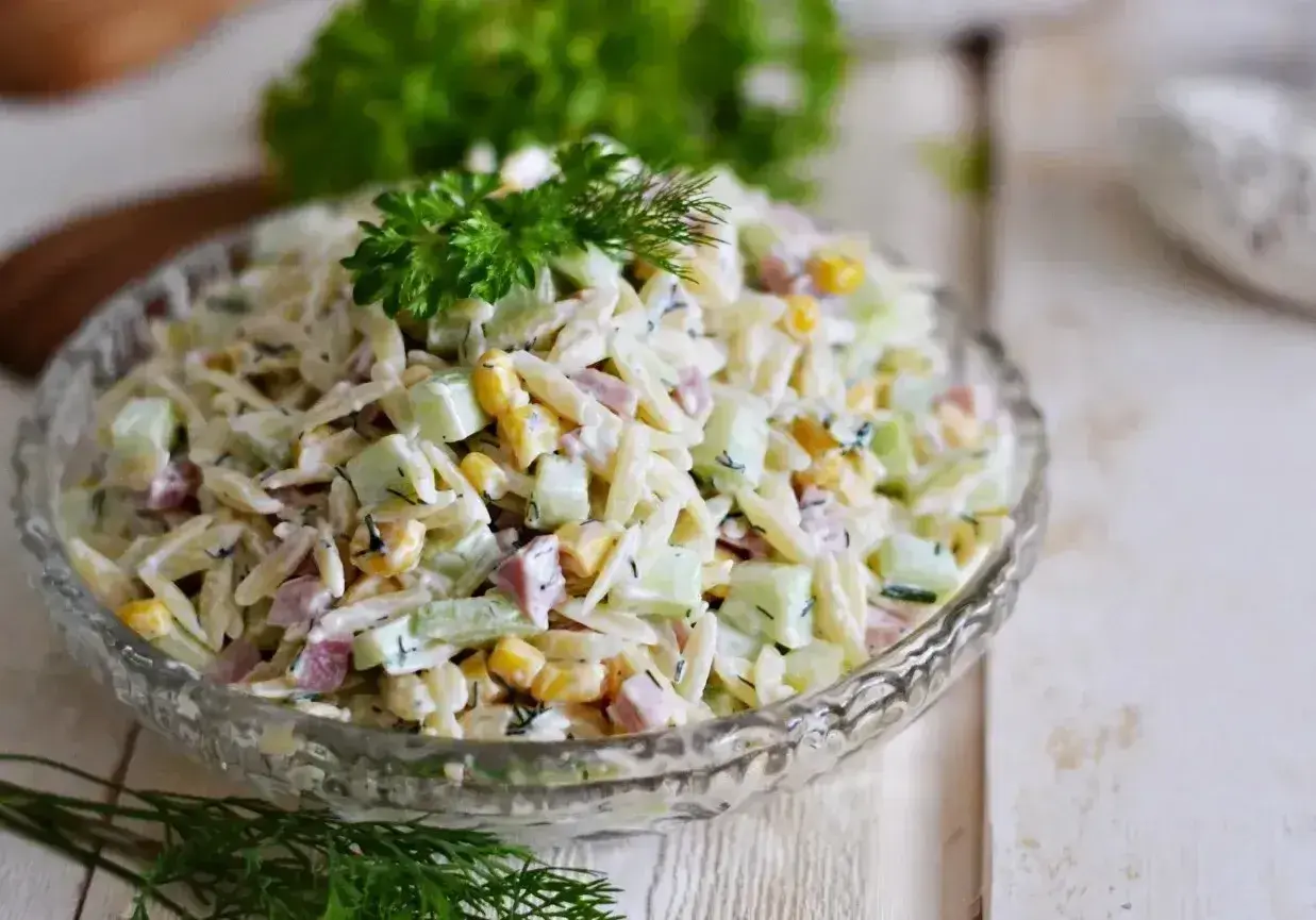 Pyszna sałatka z makaronem orzo, ogórkiem, kukurydzą i szynką, udekorowana natką pietruszki i koperkiem.