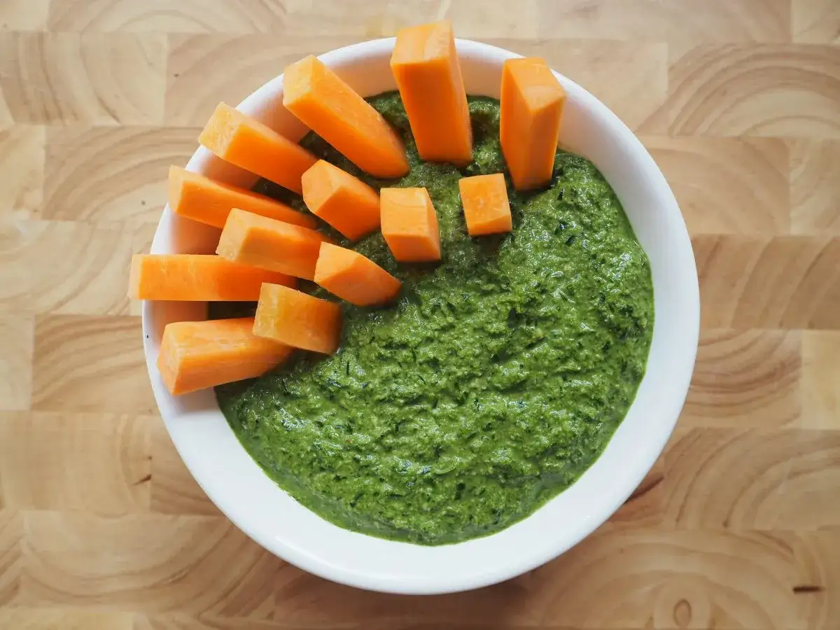 Pesto z natki marchewki podane z kawałkami marchewki w białej misce na drewnianej desce.