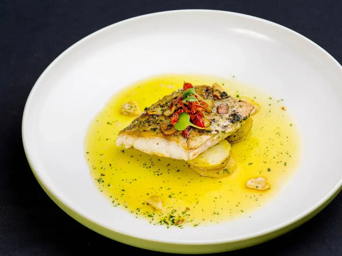 Filet ryby na plastrach ziemniaków, polany aromatycznym sosem maślano cytrynowym, ozdobiony ziołami i suszonymi pomidorami.