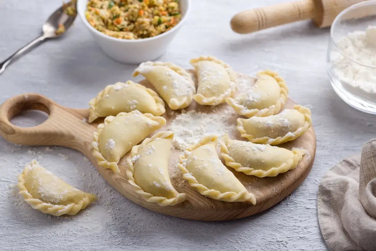 Gotowe do gotowania pierogi ułożone na drewnianej desce. W tle miska z farszem i mąka. Idealne ciasto na pierogi czeka na swoją kolej.